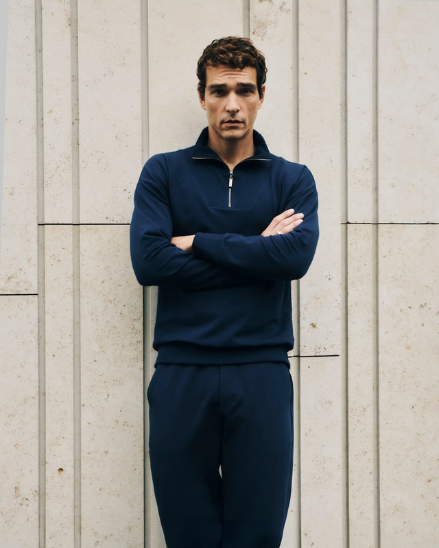 Man wearing a navy blue Natural Living tracksuit standing against a light stone wall.