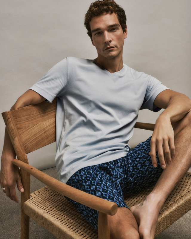 Man sitting on a wooden chair wearing a light gray t-shirt and blue patterned shorts from Night & Day.