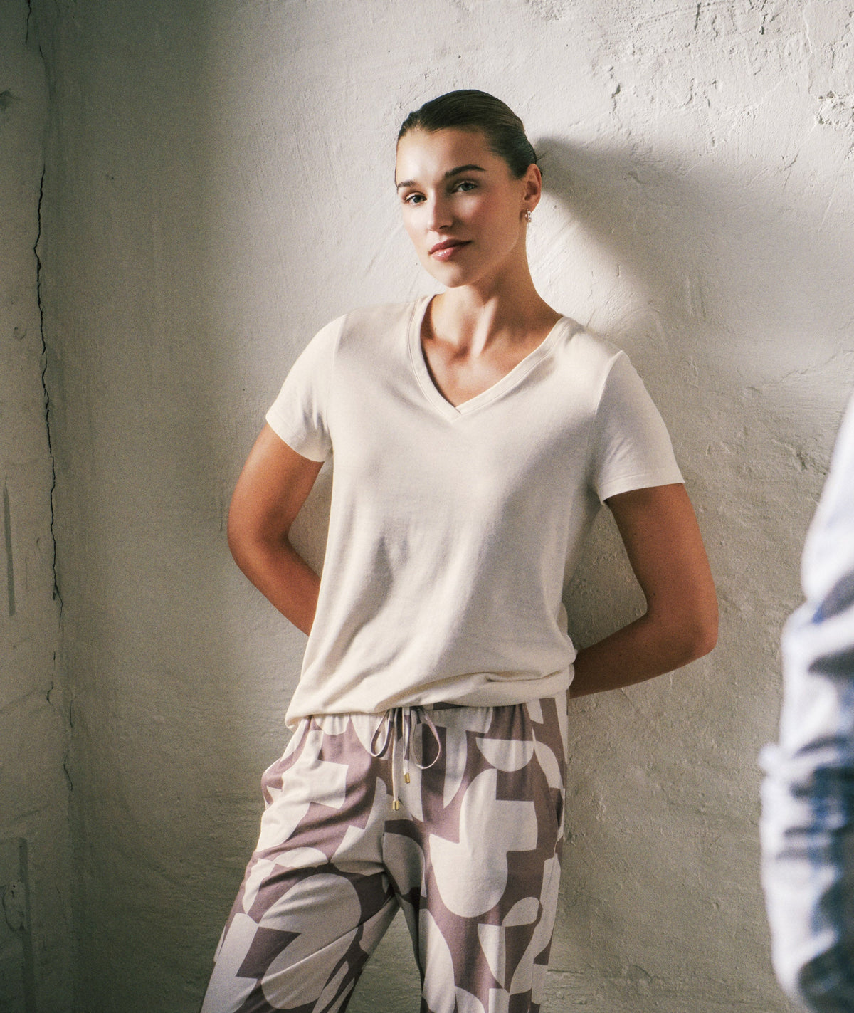 A woman stands against a textured white wall wearing a white V-neck short-sleeve top and patterned lounge pants. Another woman in a blue striped shirt is blurred in the foreground, creating a mirror-like effect.