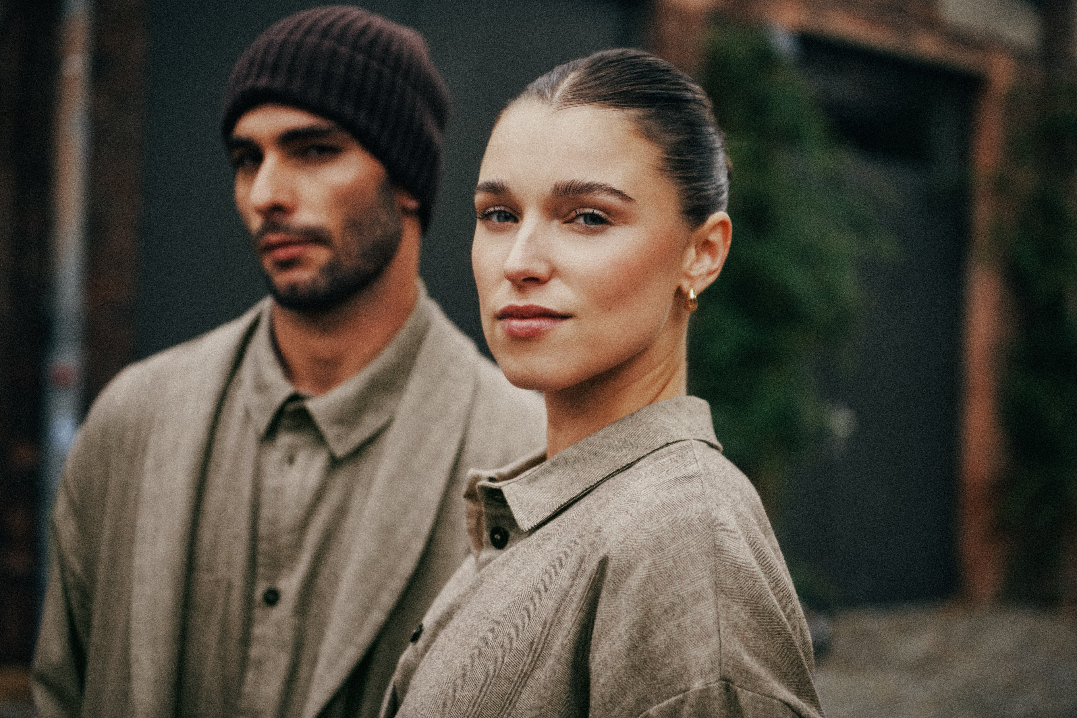 A man and a woman wearing matching muted brown and beige collared shirts and outer layers, photographed outdoors against a dark urban background.