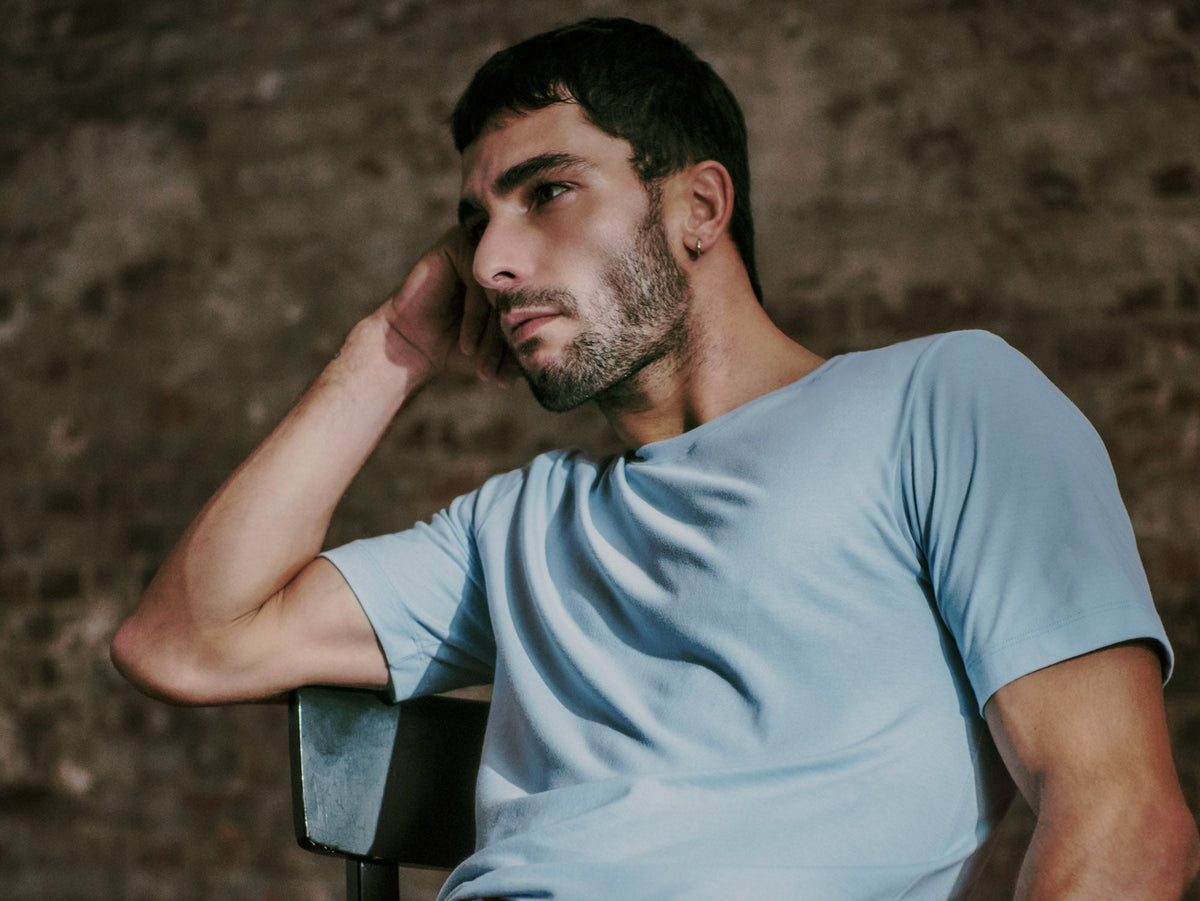 A man wearing a light blue crew-neck t-shirt sitting against a rustic textured wall.