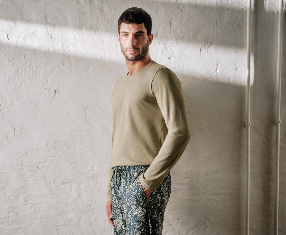 Man wearing a beige sweater and patterned pants standing against a white wall.