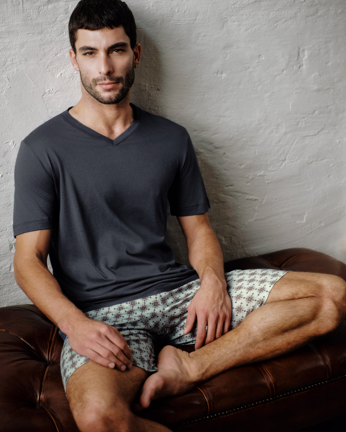 A man sitting on a brown leather couch wearing a dark gray v-neck t-shirt and patterned shorts against a textured light gray wall.