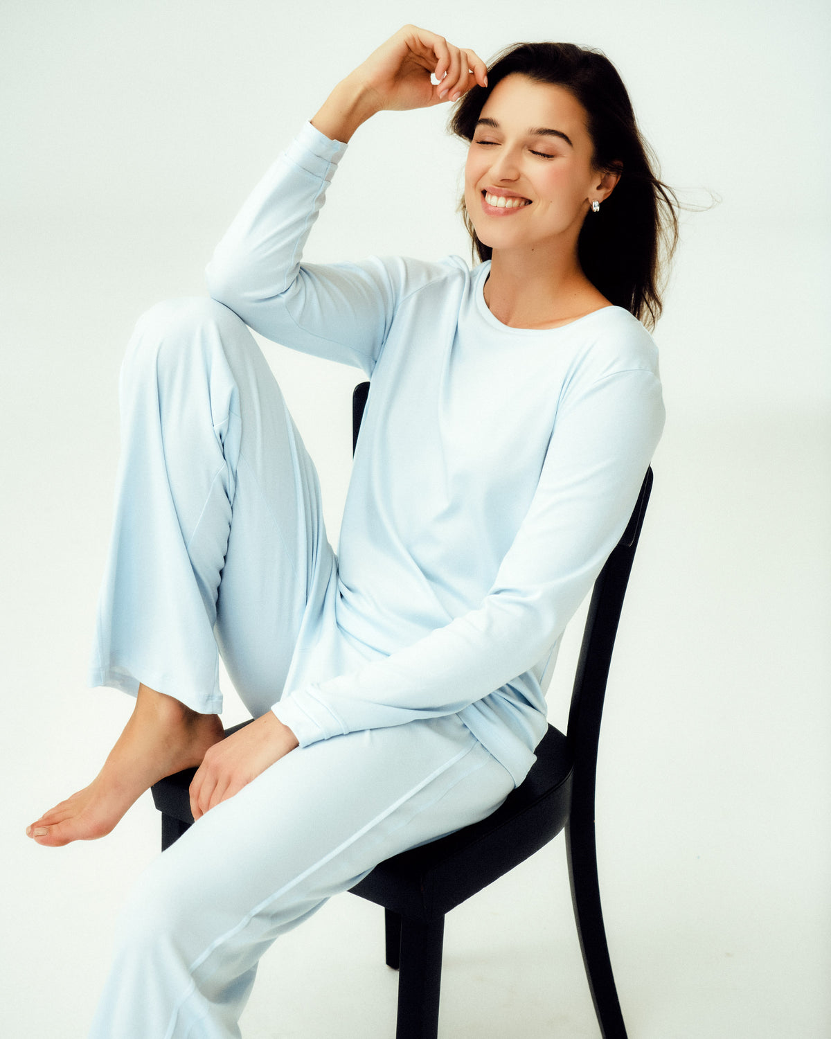 A woman wearing a light blue long-sleeve pajama set sits on a black chair against a plain white studio background.