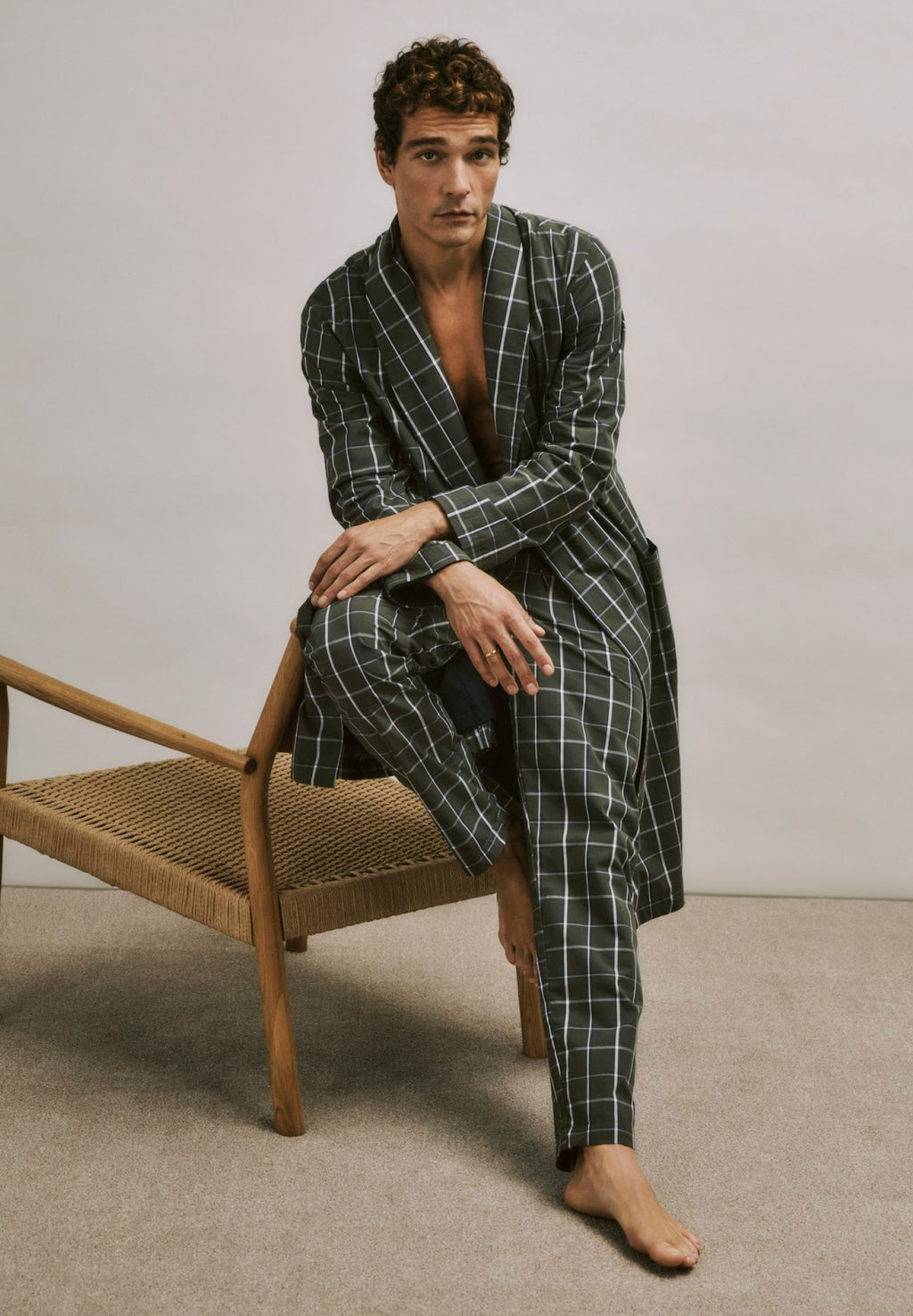 Man wearing a green checkered robe sitting on a wooden chair against a plain background