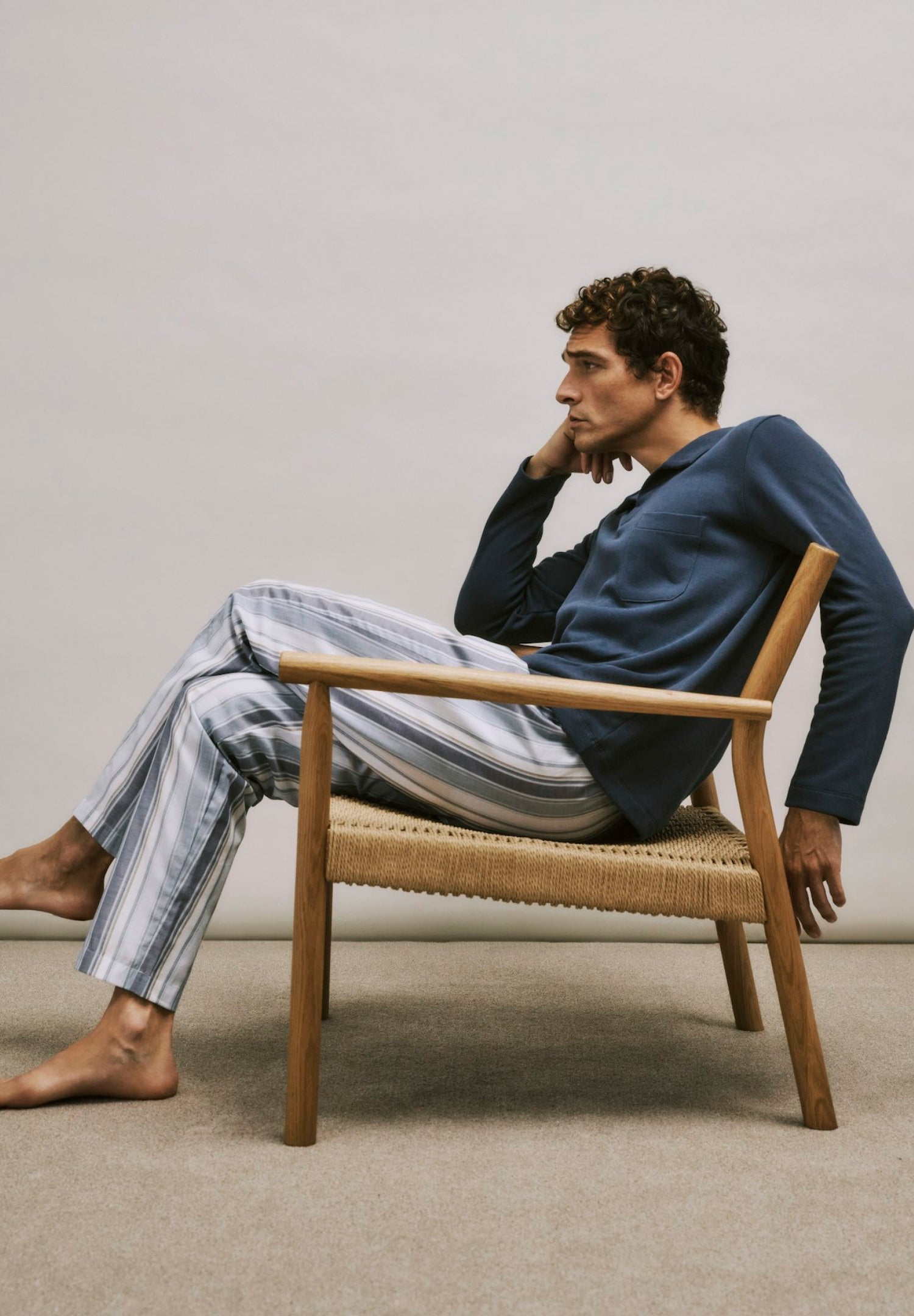 Man sitting on a wooden chair wearing a blue sweater and striped pants against a plain background