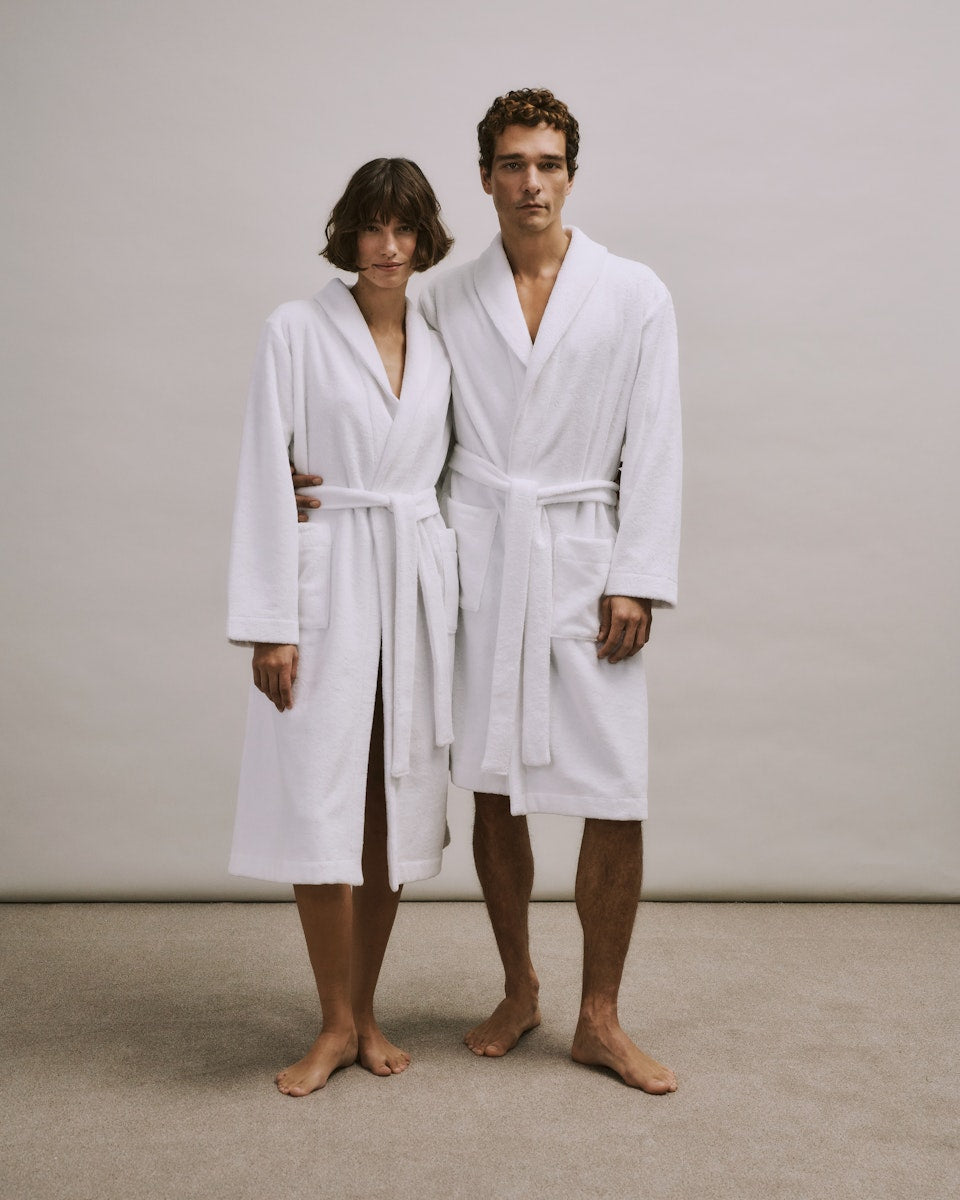 Woman and man wearing white  bathrobes from the Robe Selection Collection standing against a plain background
