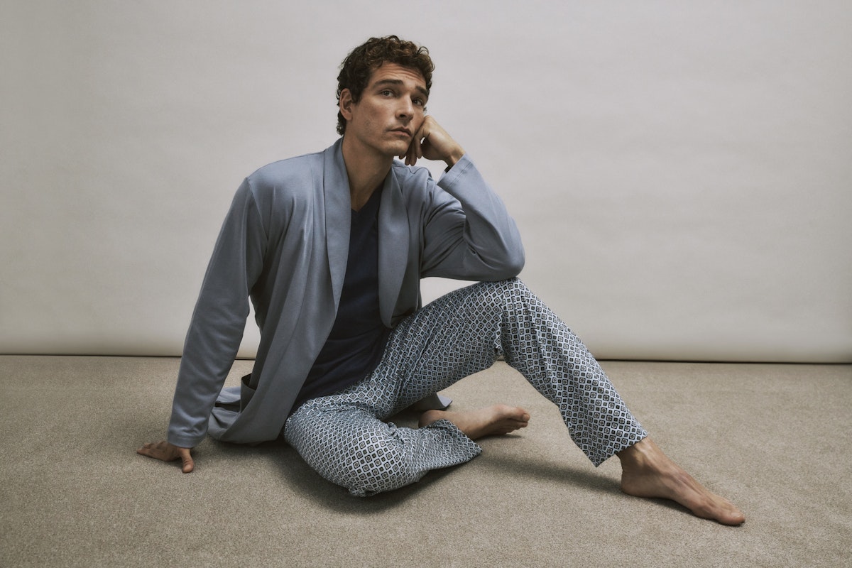 Man wearing a light blue robe and patterned pants sitting on the floor against a plain wall.