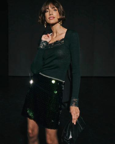 Woman wearing a dark green top with lace details and a black sequin skirt against a dark background.