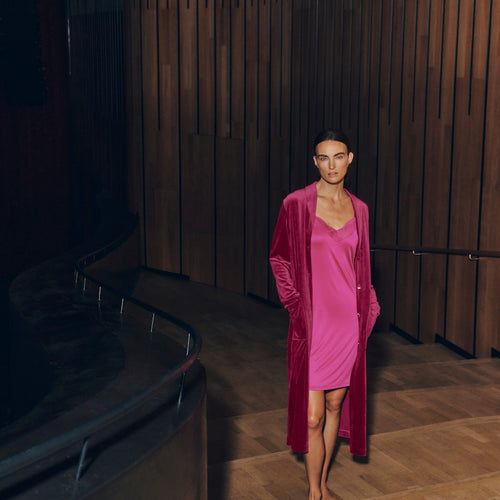 Woman wearing Lilith pink dress and Favourites robe standing in a dimly lit room with wooden walls.