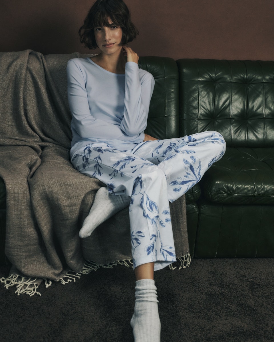 Woman sitting on a dark green leather couch wearing a powder blue top and white pants with blue floral pattern.