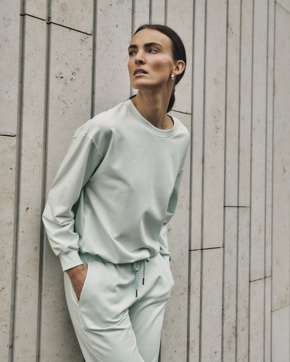 Woman wearing a light green sweatshirt and pants against a textured wall.