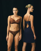 Two models standing against a black studio background. The model on the left wears a brown lace bra and matching lace underwear, while the model on the right wears a dark navy blue sleeveless dress with a back cutout.