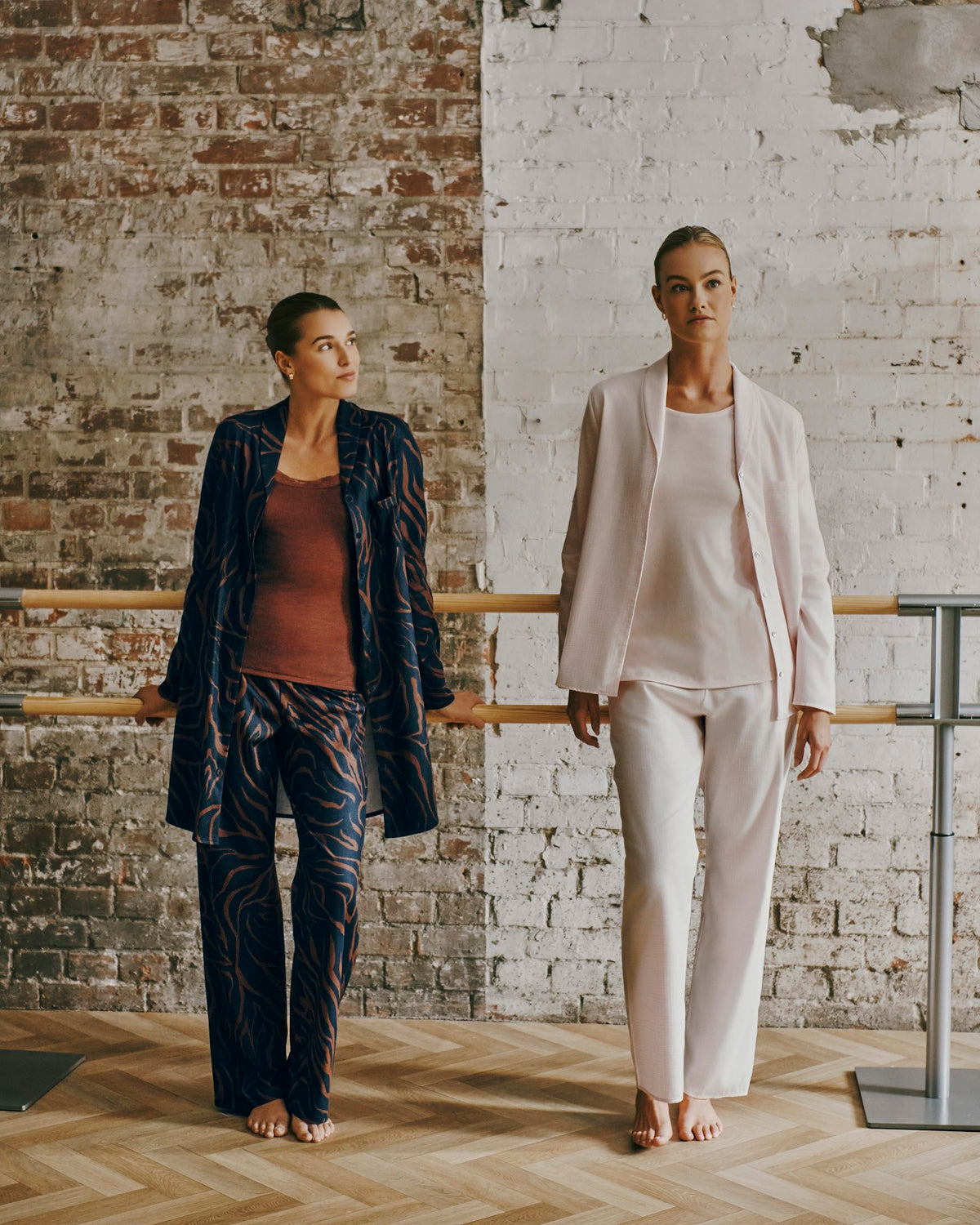 Two models standing in a studio with a brick and white plaster wall background, wearing matching lounge sets. The model on the left wears a dark navy patterned blazer and trousers over a rust-brown top.