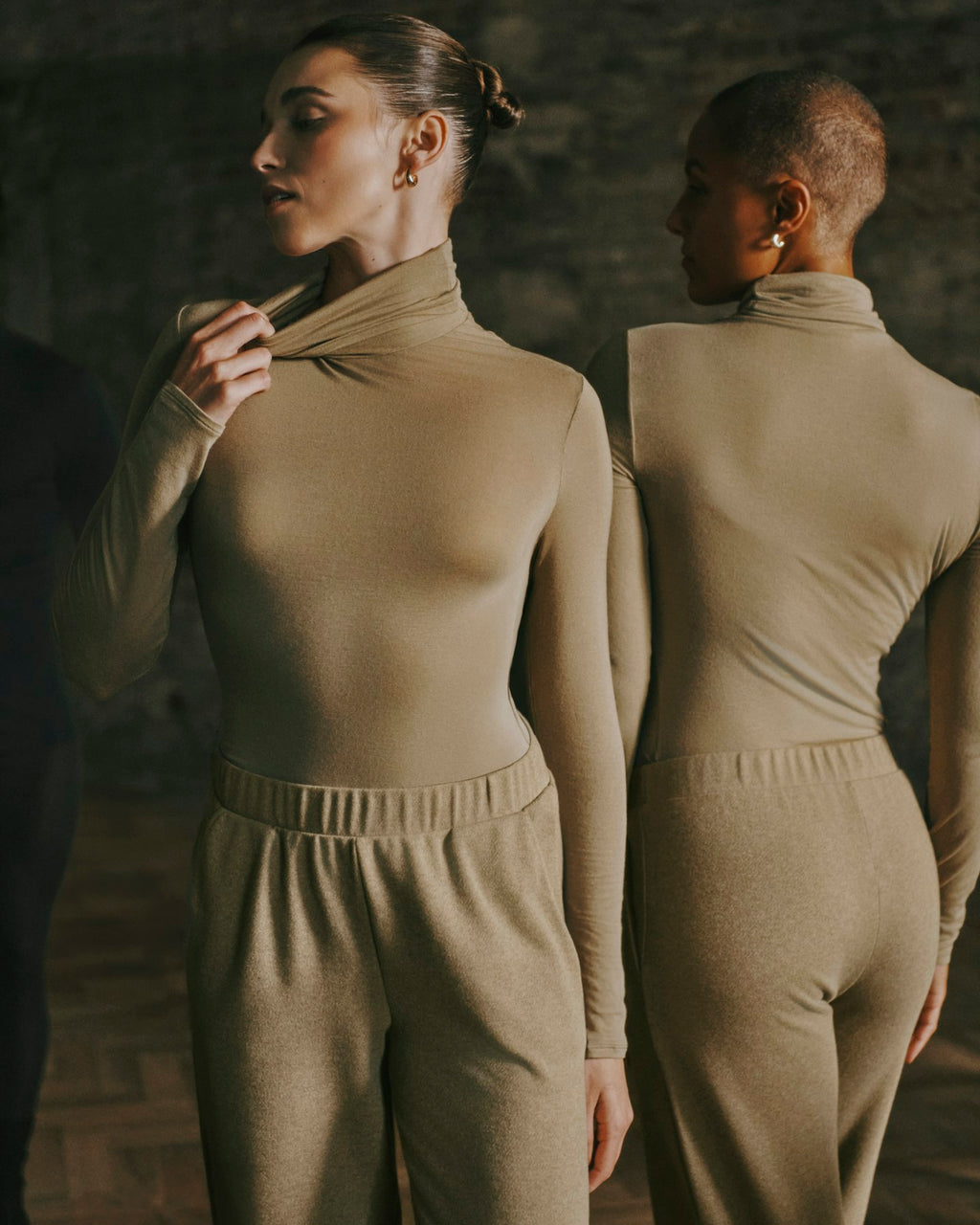 Two models wear a matching beige knit mock-neck top and jogger pants set, shown from both front and back angles against a dark rustic brick background.