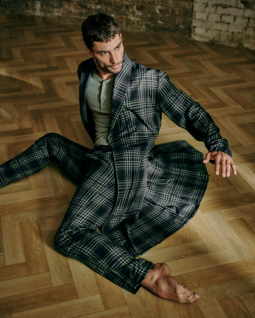 A man sits barefoot on a wooden herringbone floor wearing a dark gray and black plaid lounge set with a matching robe and matching pants.
