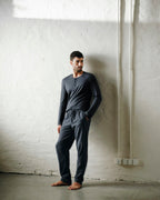 A man wearing a dark gray long-sleeve henley shirt and matching dark gray sweatpants stands barefoot against a textured white plaster wall.