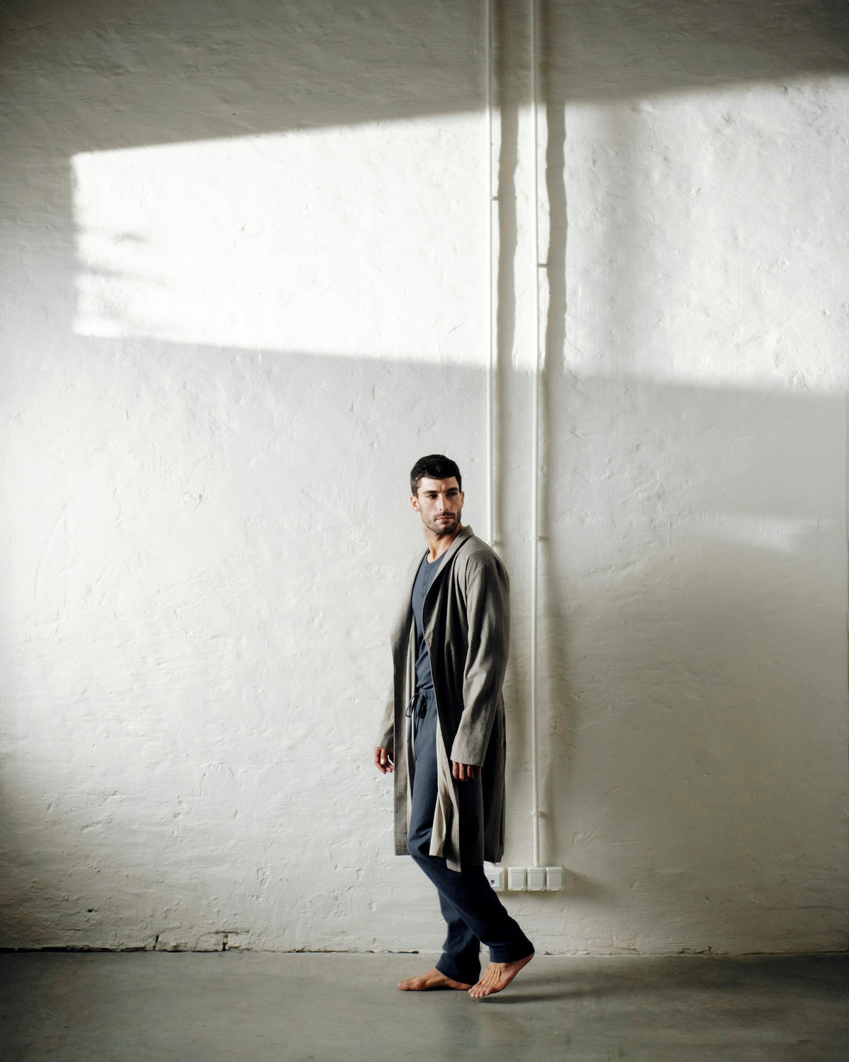 A man stands barefoot against a textured white wall in a minimalist room, wearing a long gray robe over a shirt and dark trousers.