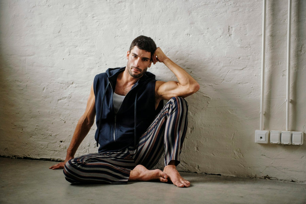 A man sits barefoot against a textured white wall, wearing a navy sleeveless zip-up hoodie over a white tank top and striped pajama pants in black, gray, and brown.