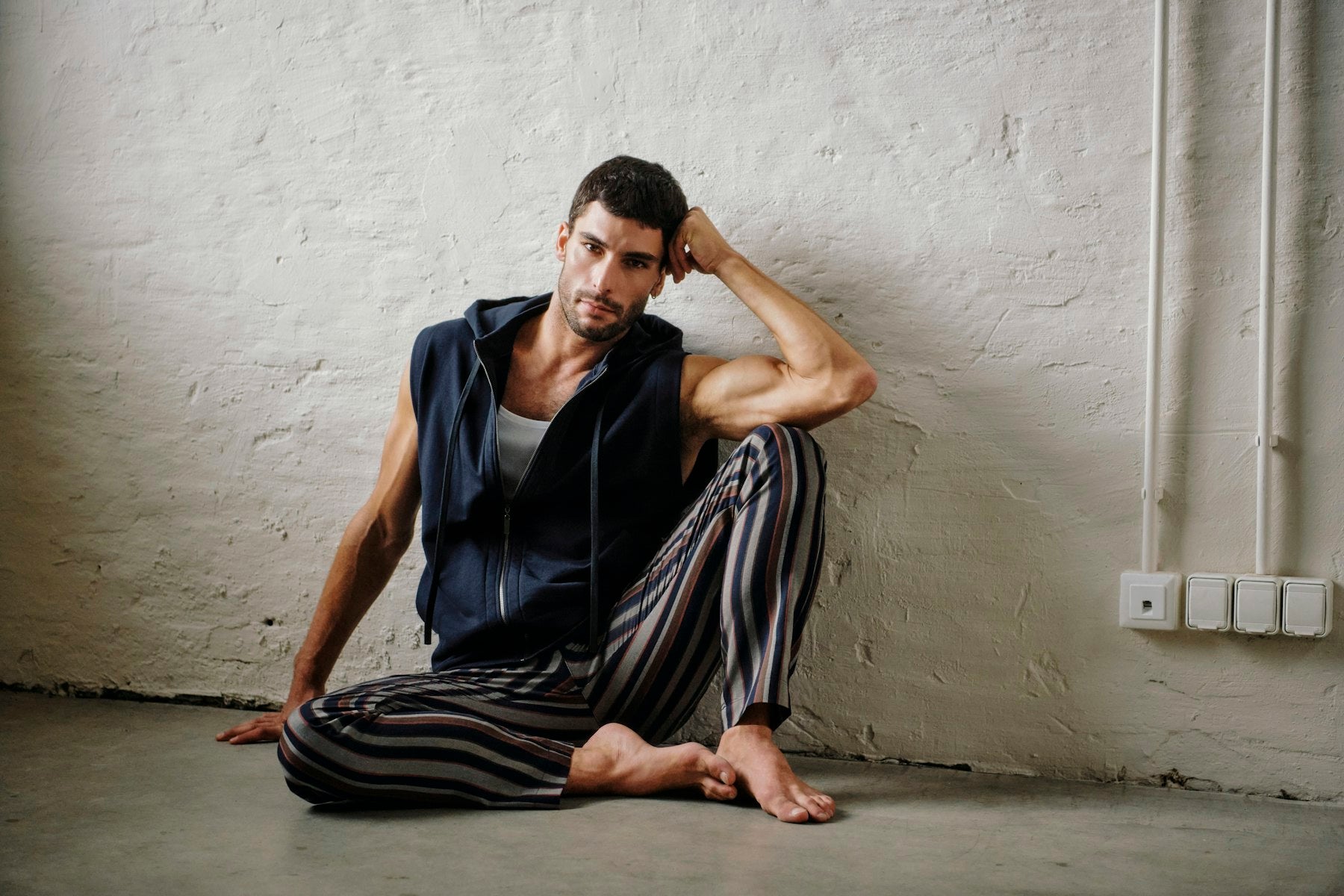 A man sits barefoot against a textured white wall, wearing a navy sleeveless zip-up hoodie over a white tank top and striped pajama pants in black, gray, and brown.