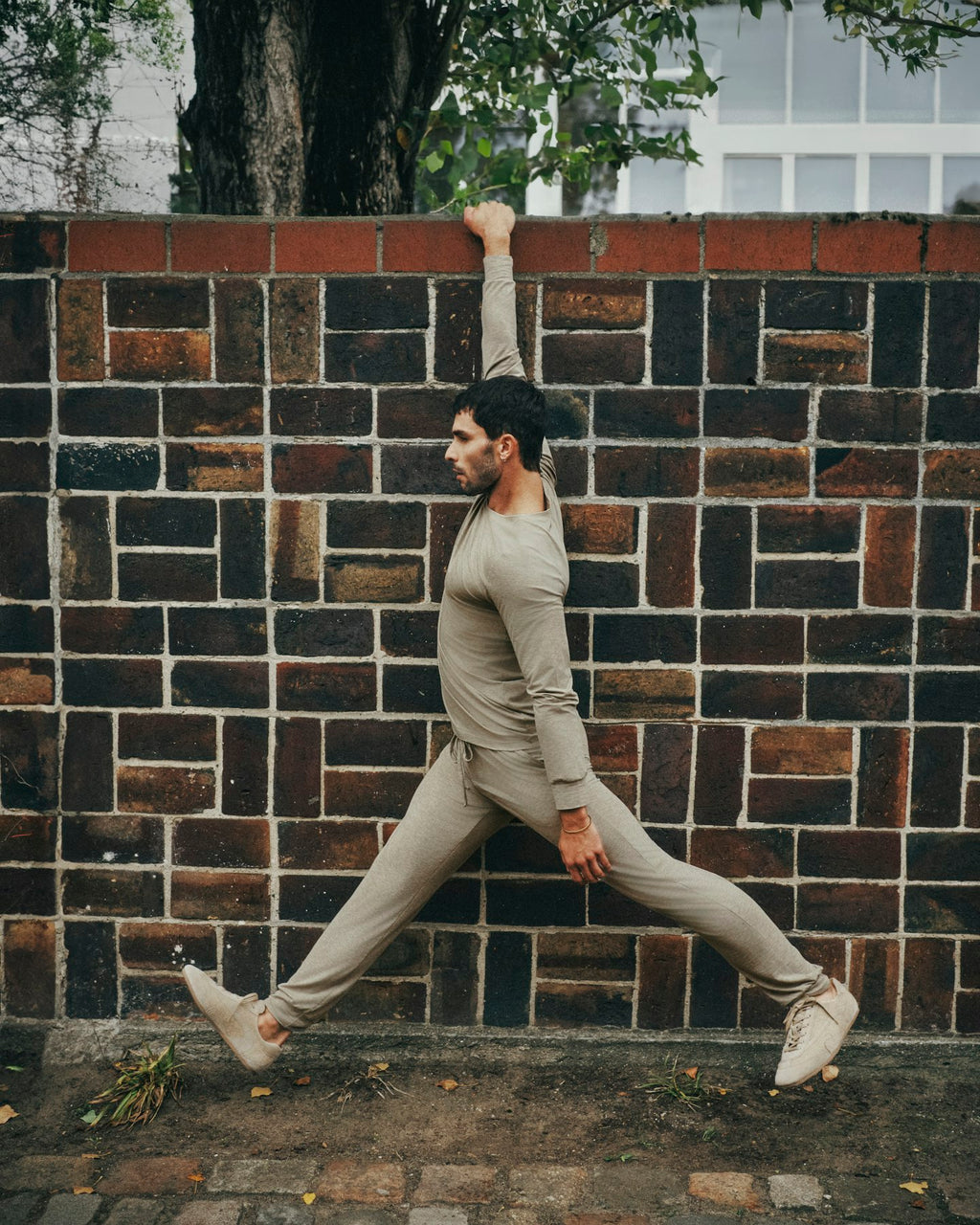A man in a beige long-sleeve athletic shirt and matching jogger pants stretches against a dark brick wall outdoors.