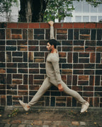 A man in a beige long-sleeve athletic shirt and matching jogger pants stretches against a dark brick wall outdoors.