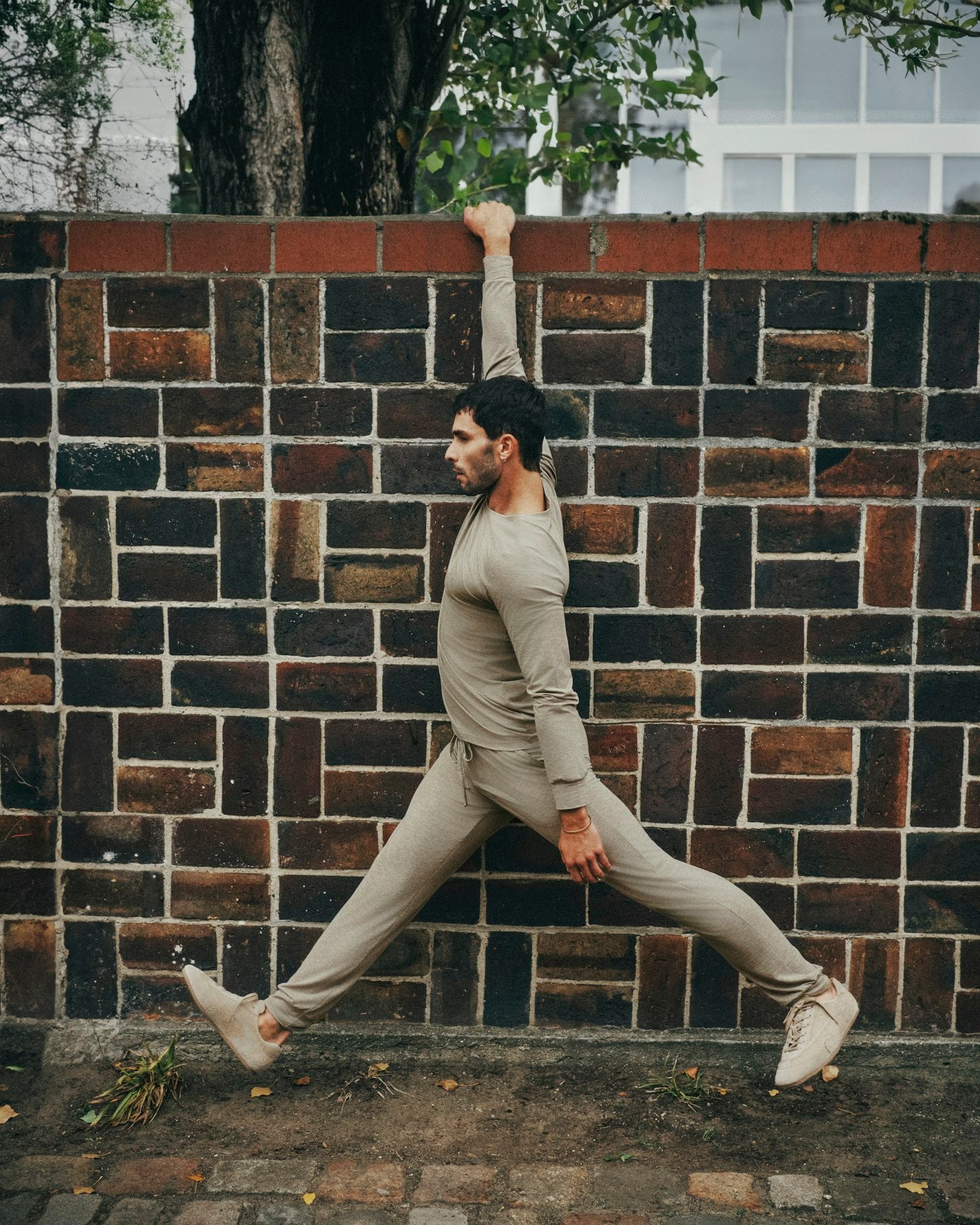 A man in a beige long-sleeve athletic shirt and matching jogger pants stretches against a dark brick wall outdoors.