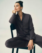 A woman wearing a dark charcoal gray pajama set with a button-front long-sleeve top and matching pants, seated on a black chair against a light gray studio background.