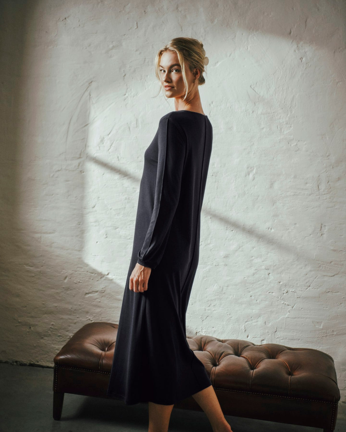 A woman wearing a long-sleeve black midi dress stands in profile next to a brown tufted leather bench against a white textured wall with dramatic sunlight and shadows.