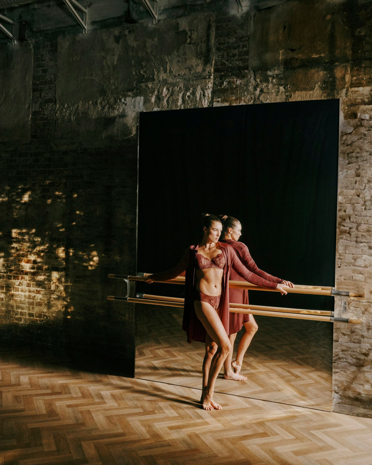 A female dancer in matching burgundy and brown dancewear pose in front of a large black mirror in a rustic studio with brick walls and wooden floors.