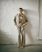 A model stands barefoot against a textured white plaster wall, wearing a matching beige two-piece outfit consisting of zip-up jacket and wide-leg trousers.