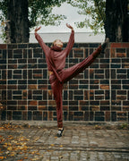 A person in a matching rust-brown tracksuit poses dynamically with one leg extended upward in front of a dark brick wall outdoors.