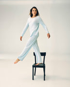 A woman modeling a light blue long-sleeve pajama set, standing barefoot on a black chair in a studio setting.