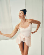 A woman modeling a pale blush pink lingerie set consisting of a camisole top with lace trim and a matching sheer wrap skirt, posed against a bright white studio backdrop.