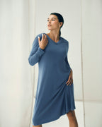 A woman modeling a long-sleeve, V-neck, knee-length dress in a dusty slate blue color, standing against a soft, off-white studio backdrop.