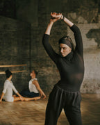 A woman in a black long-sleeve mock neck top and black sweatpants stretches her arms overhead in a rustic studio with brick walls and wood floors. Two other dancers are seated in the background.
