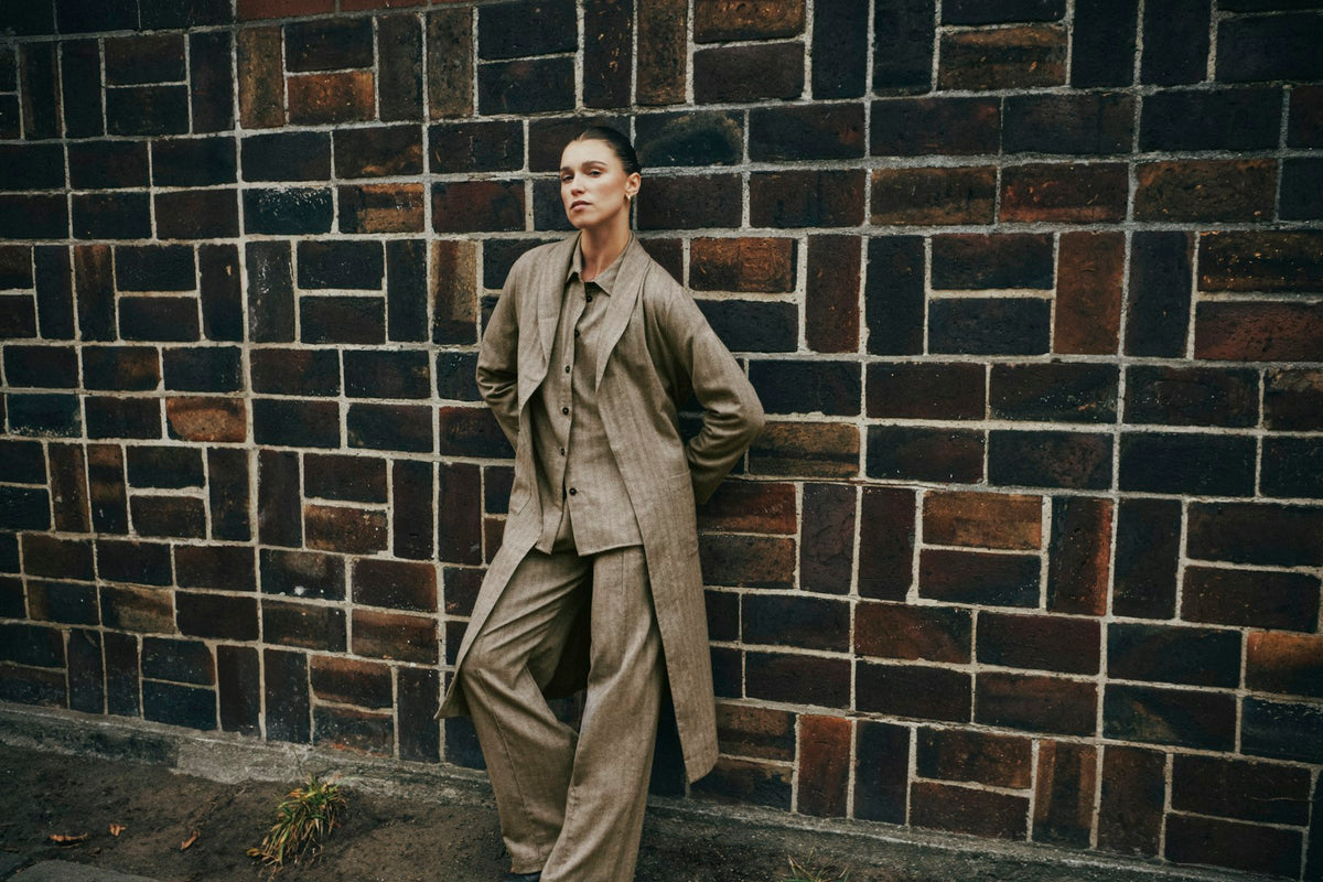 A model wearing a matching beige pinstripe suit and long overcoat leans against a dark brick wall.
