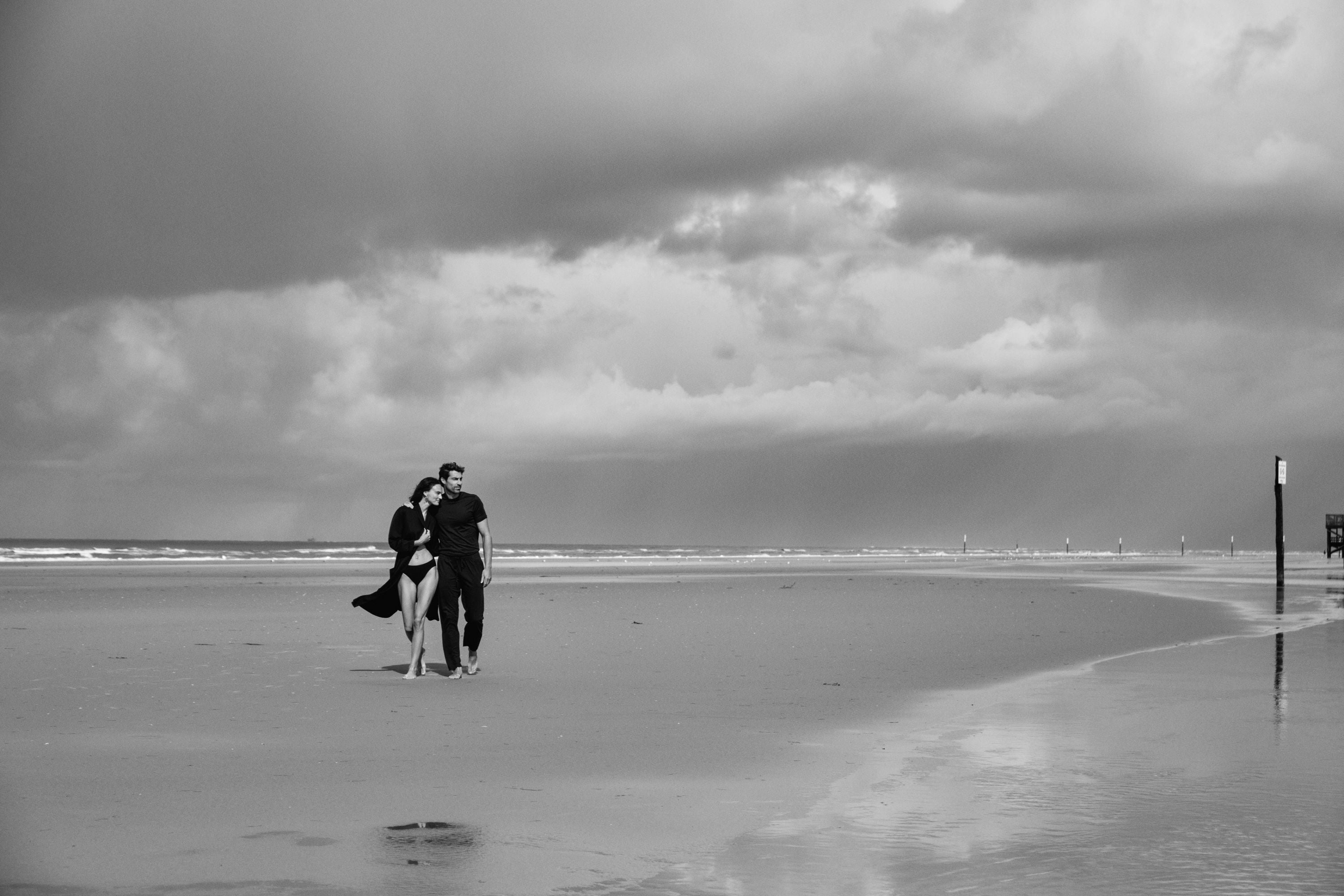 Black and white photo of a couple walking on a beach with a stormy sky.