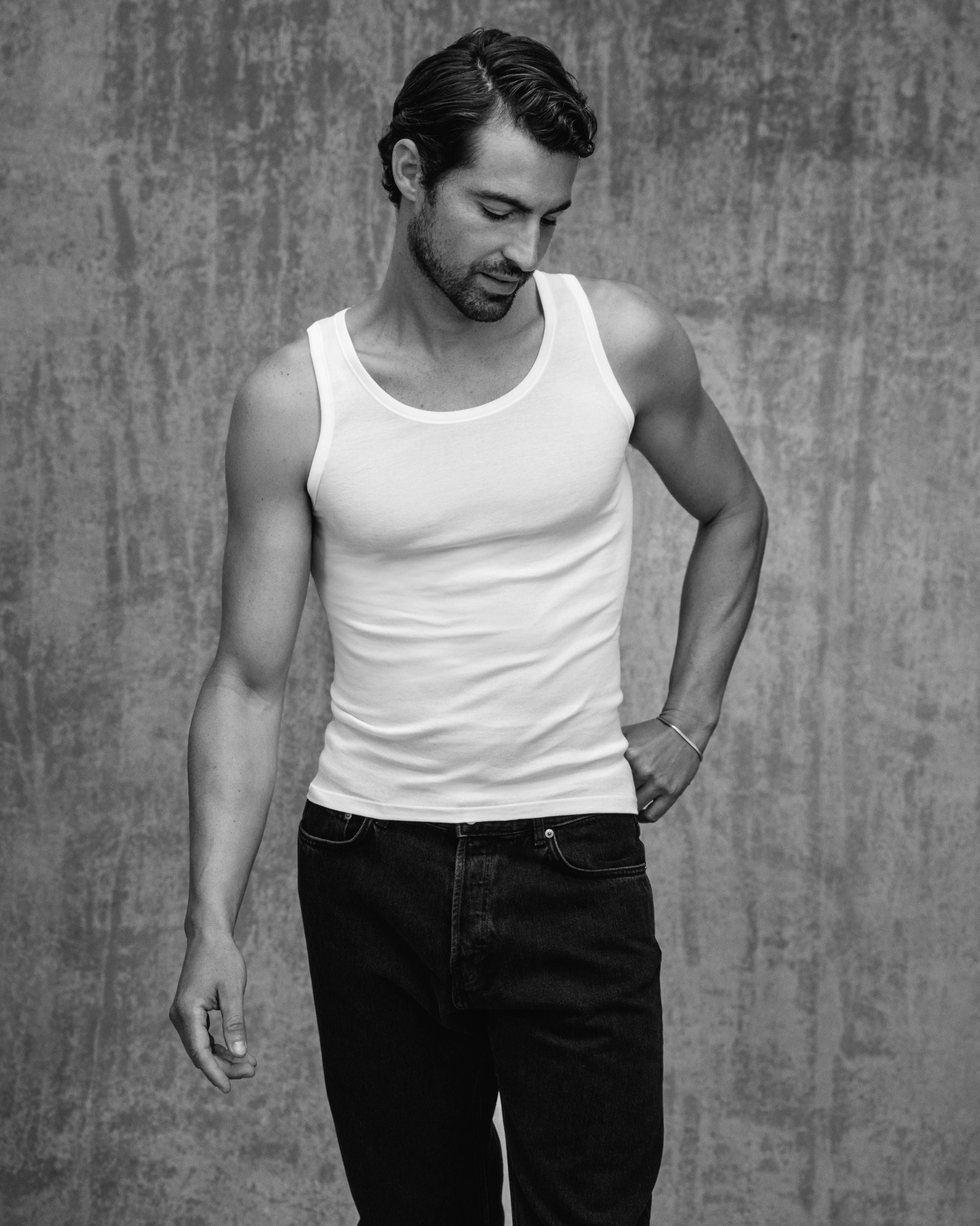 A man wearing a fitted white ribbed tank top and dark jeans, standing against a textured gray wall in a black-and-white portrait-style photo.