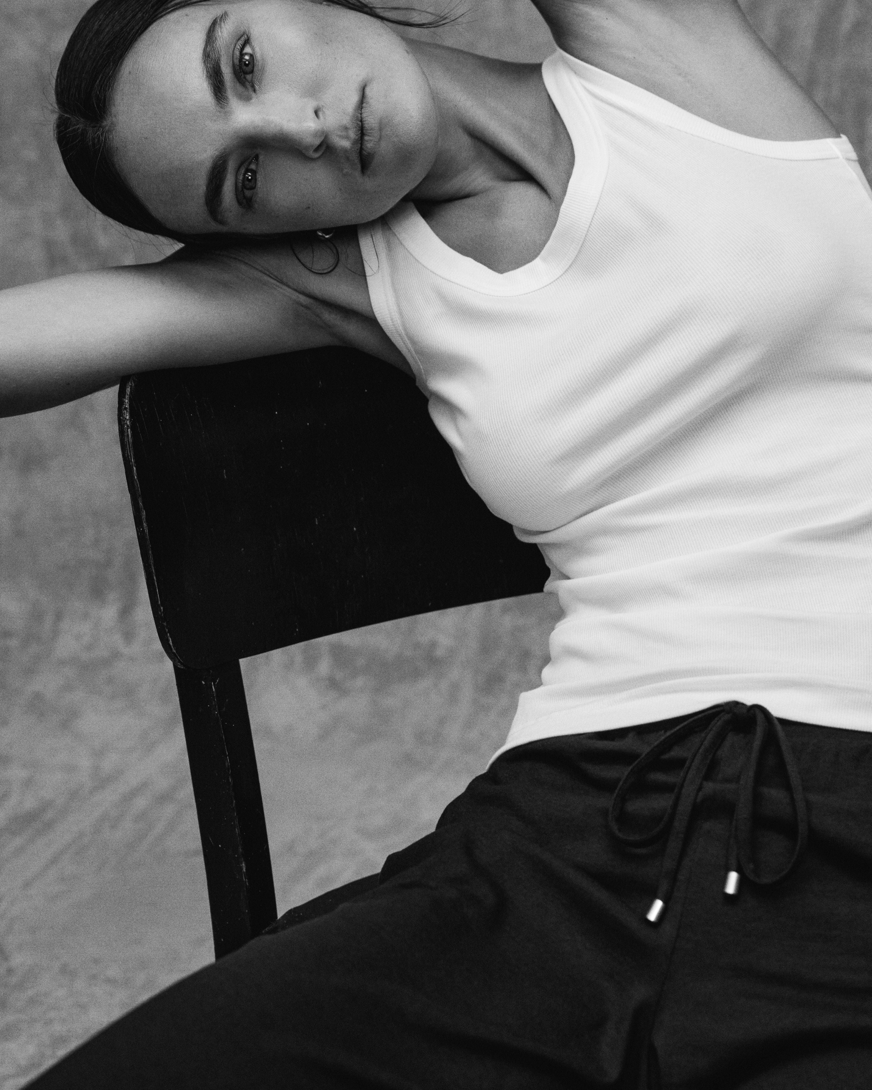A black and white editorial photograph of a woman leaning against a dark chair, wearing a white ribbed tank top and dark drawstring sweatpants.