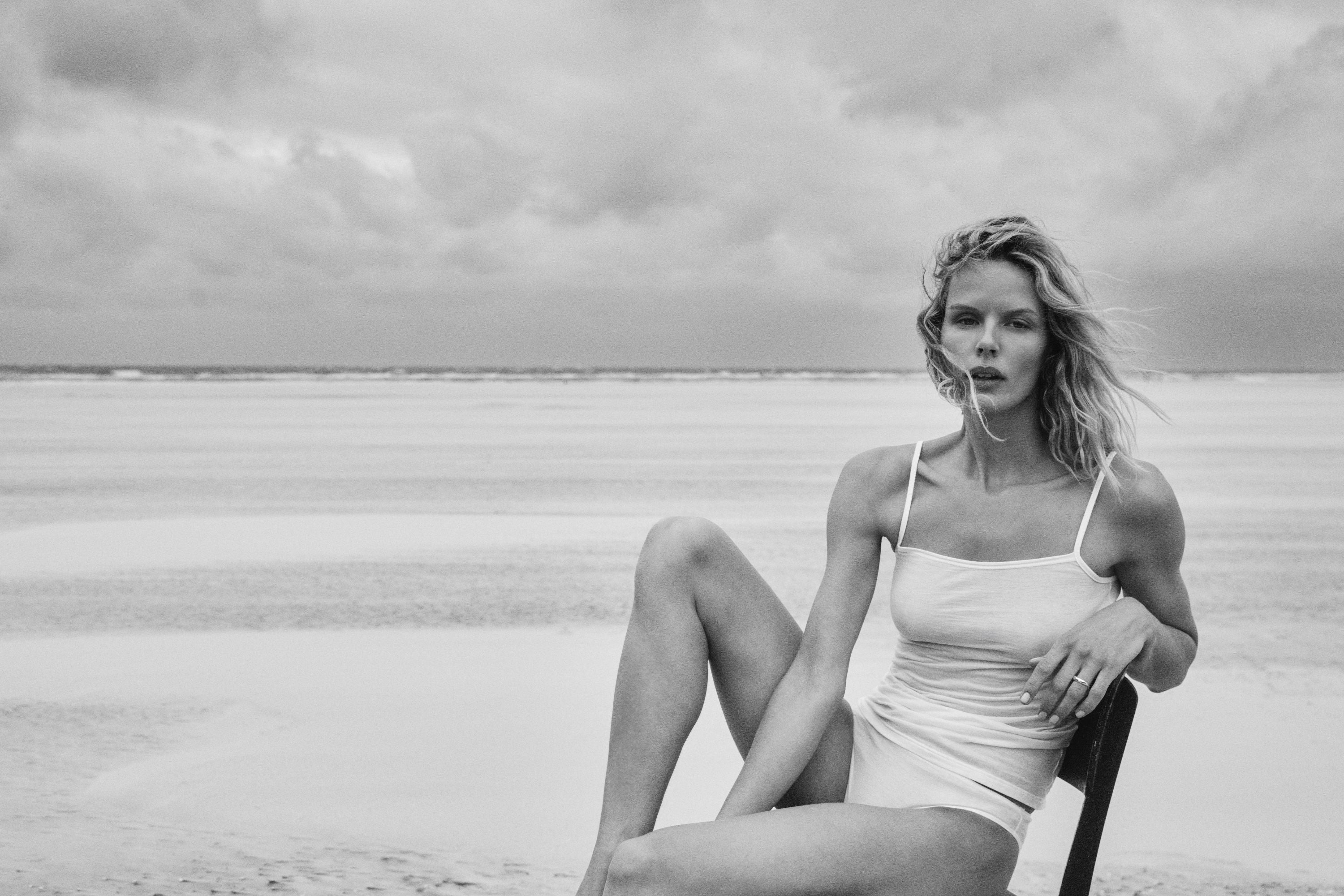 A black and white fashion photograph of a woman sitting on a beach chair on a sandy beach wearing a white camisole and panties