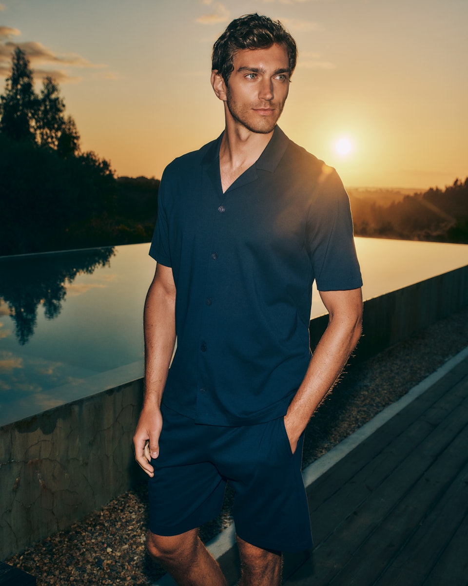 Man standing on deck at sunset with his hand in this pocket wearing Night And Day Notch Collar Button Front Cotton Shirt in Deep Navy, paired with Night And Day Cotton Drawstring Shorts