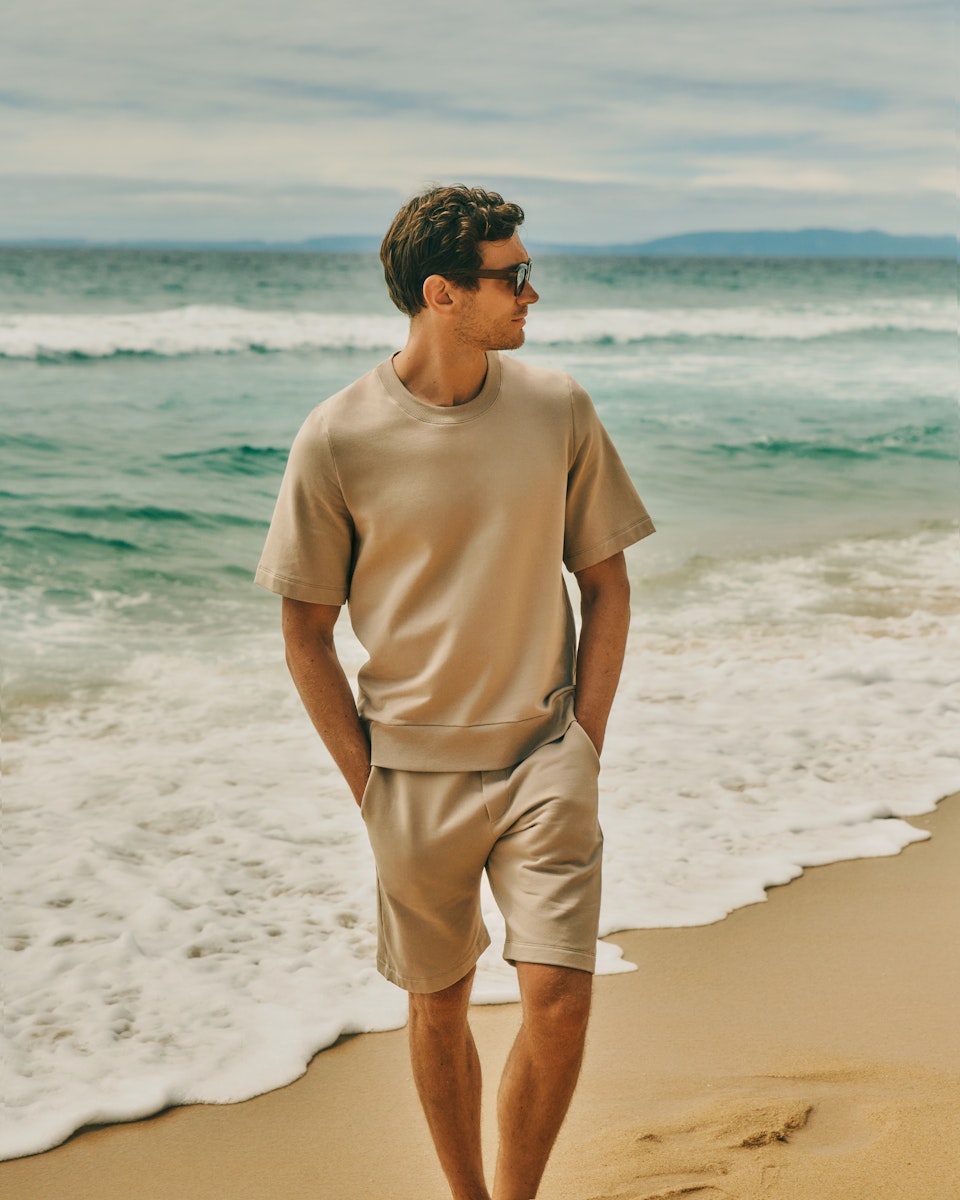 Man walking on the beach with his hands in his pockets, wearing Natural Living Organic Cotton Sweatshirt in Savanna and Natural Living Organic Cotton Drawstring Shorts 
