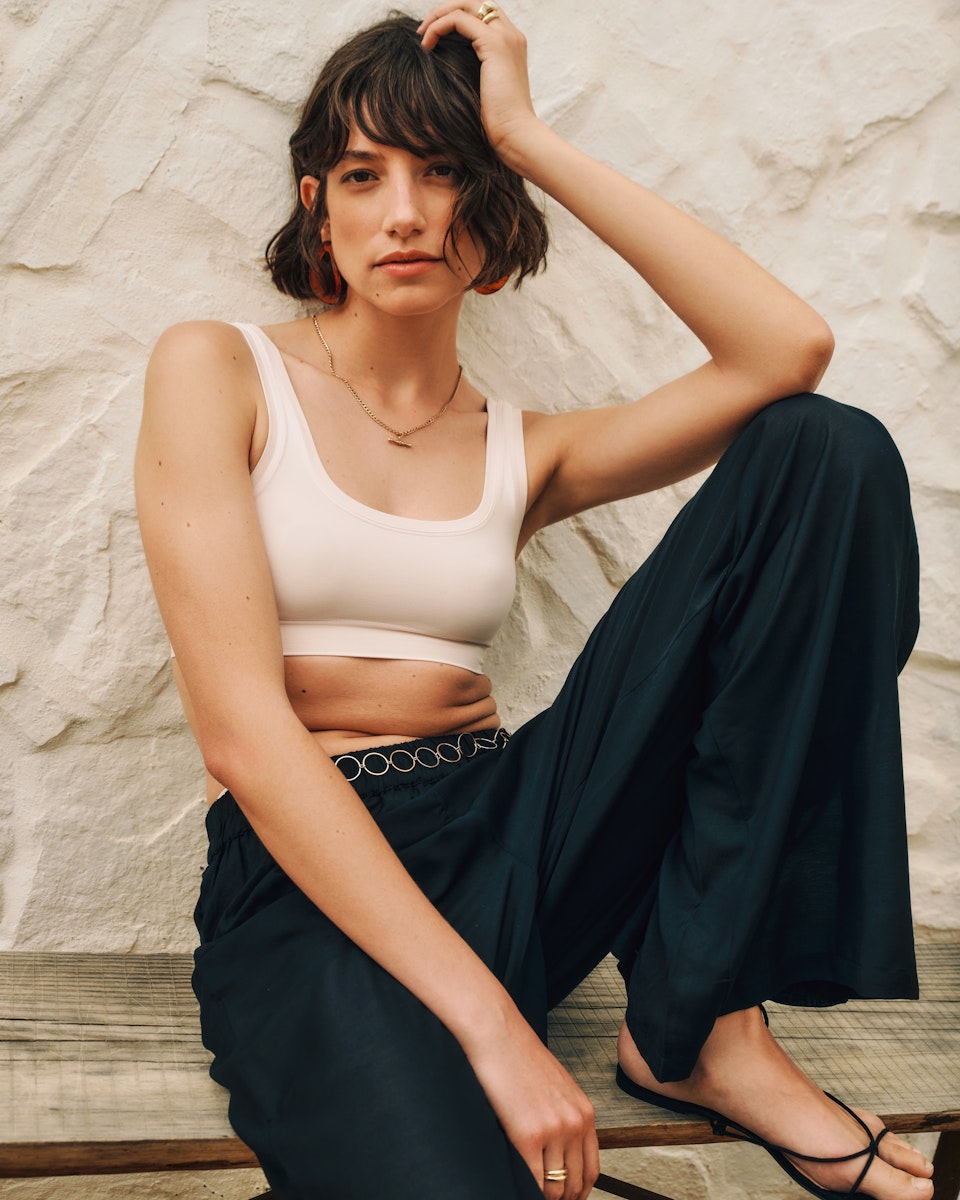 Woman sitting on a wooden bench wearing a white tank top and dark pants against a textured wall.