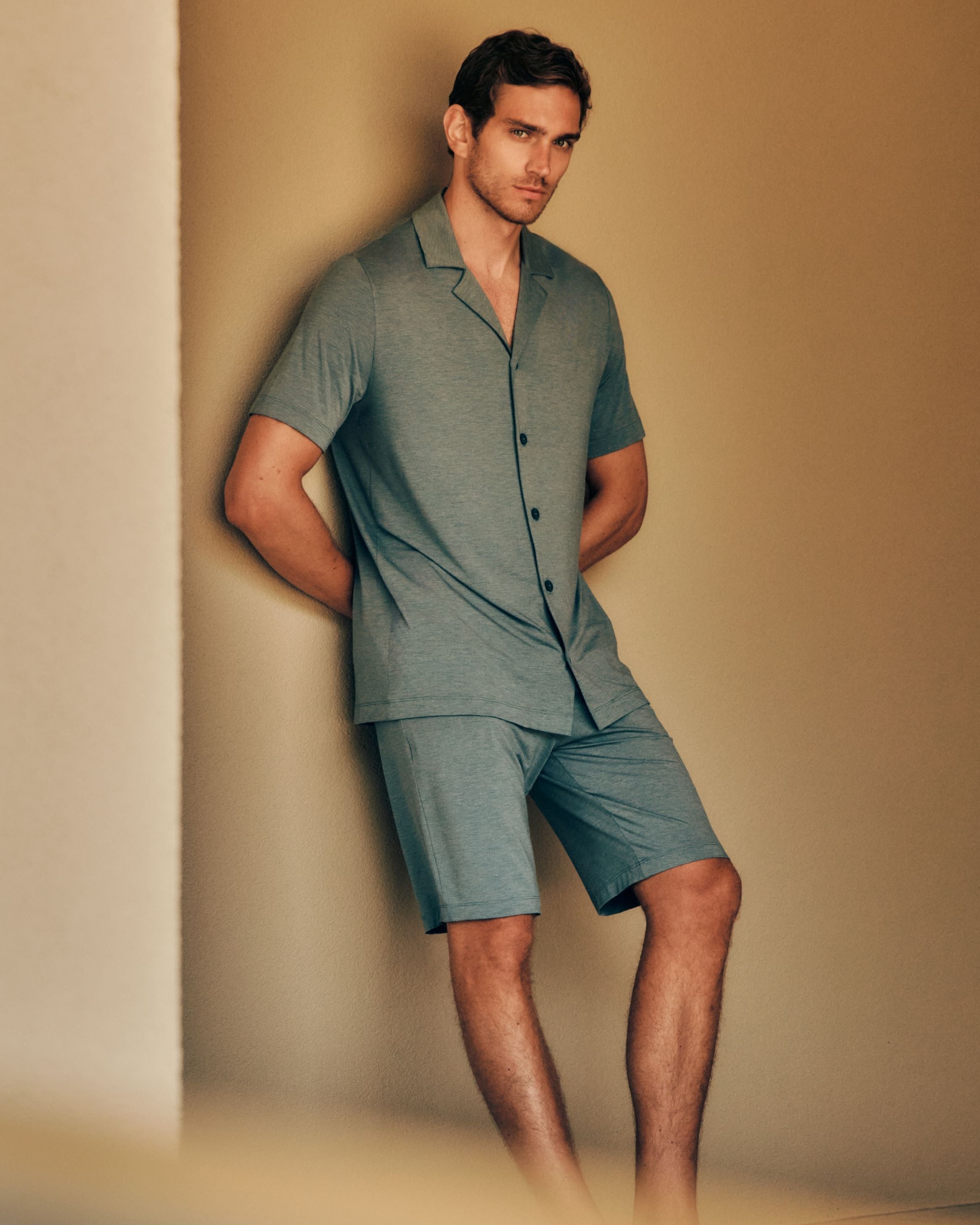 Man leaning against a wall with his hands behind his back, wearing the Casuals Button Front Shirt and Shorts in Ocean Mélange.