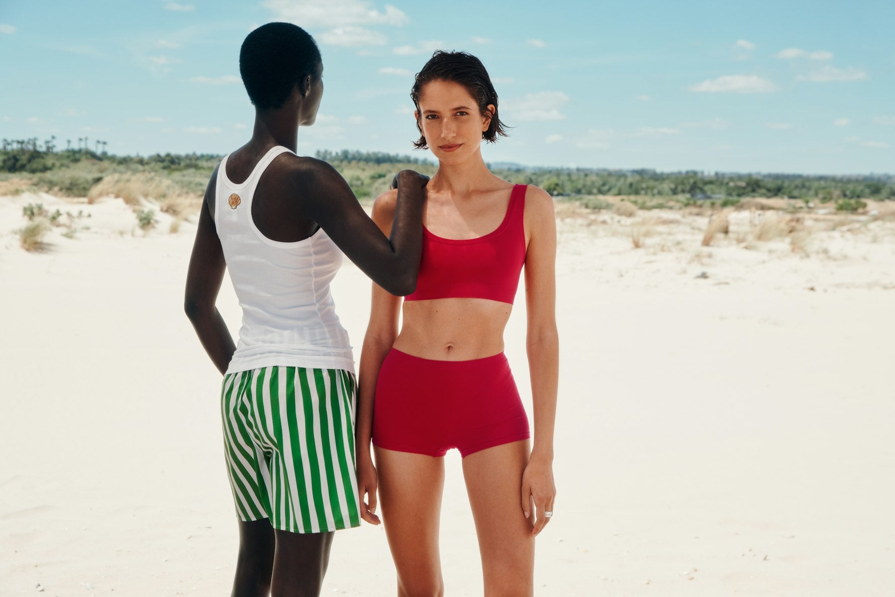 Two women standing on the beach  wearing a red outfit and white tank top with green and white striped shorts