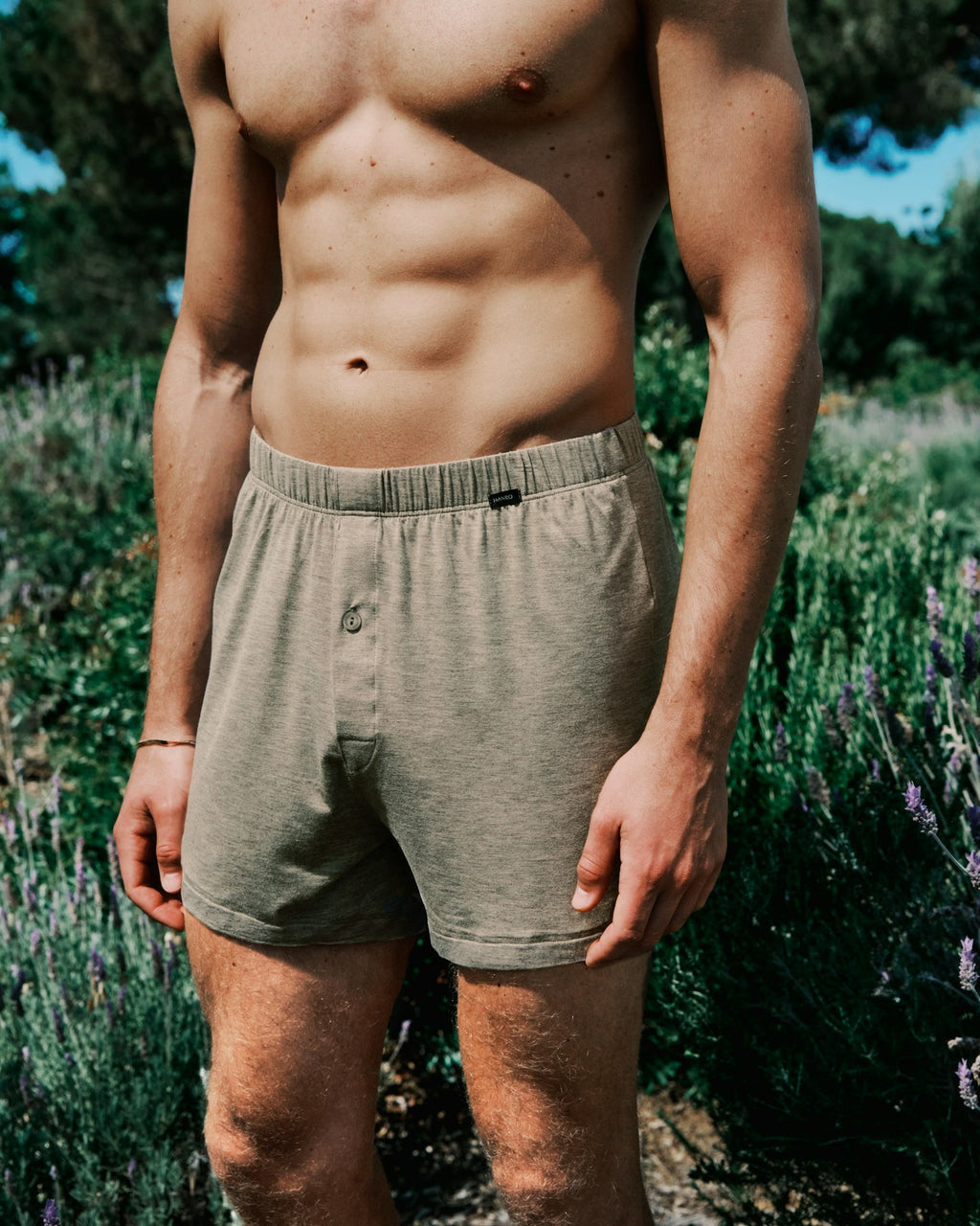 Man wearing brown boxer briefs standing in a natural setting with greenery.
