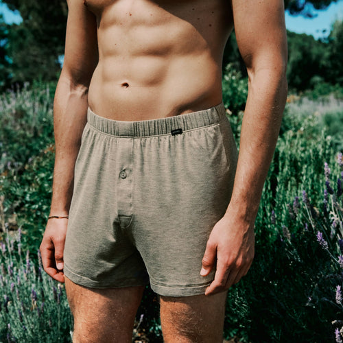 Man wearing brown boxer briefs standing in a natural setting with greenery.