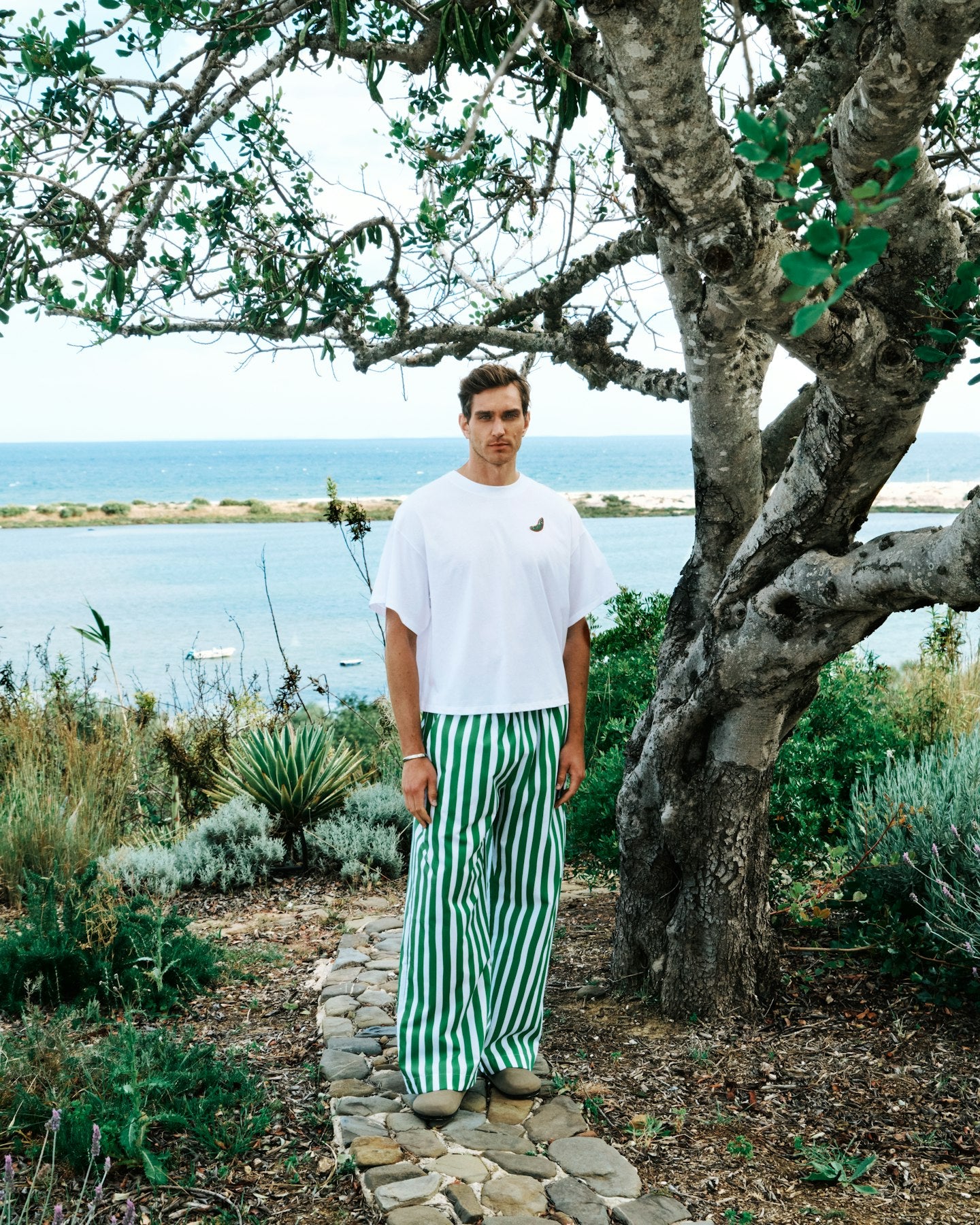 Man standing on a stone path with ocean view, wearing a white t-shirt and green striped pants.
