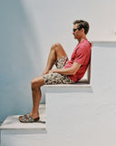 Man sitting on a white ledge wearing a red shirt and patterned shorts.